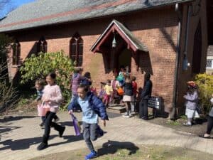Children joyfully run outside a brick building with a pointed entrance. A person observes nearby. A