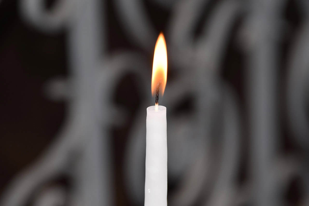 A lit white candle with a gentle flame against a blurred, ornate background, creating a serene and contemplative atmosphere.