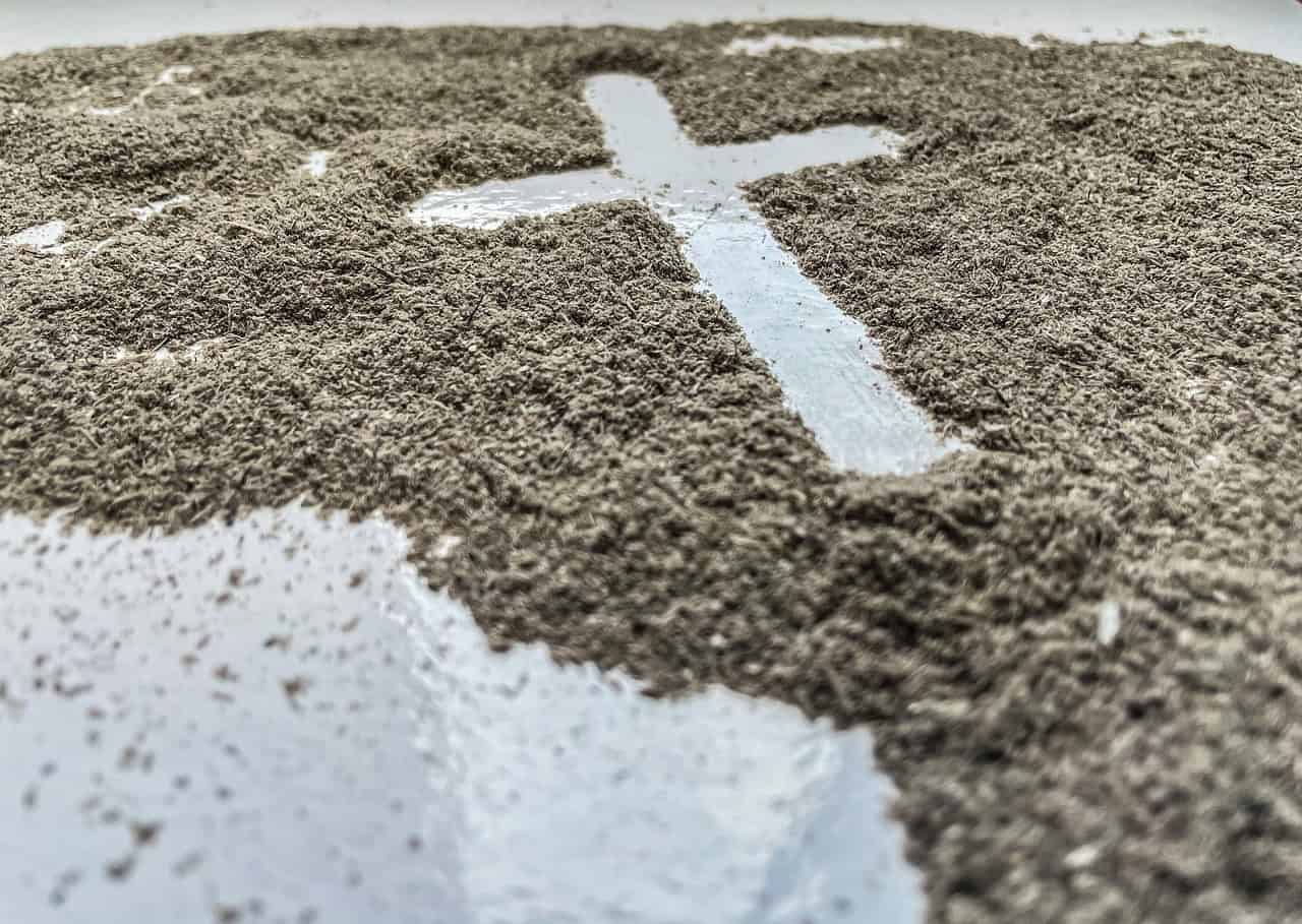 A cross-shaped space is visible in scattered gray ash on a reflective white surface, symbolizing reflection or remembrance.